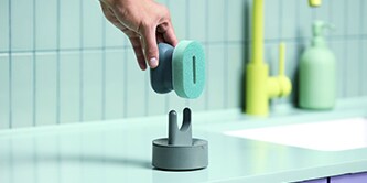 Main posant une brosse main vaisselle Brite de Scotch-Brite™ sur un porte-éponge posé sur le plan de travail de la cuisine à côté de l’évier.
